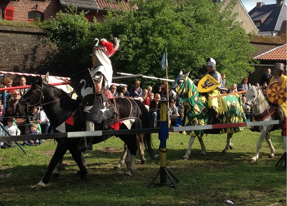 Ritterspiele im Schloss Schkopau. 2 Ritter reiten auf Pferden mit mittelalterlichen Rüstungen auf einer WIese. Dahinter steht Publikum.